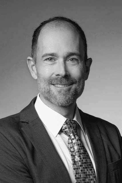Black and white portrait of a smiling man in a suit and patterned tie against a plain background.